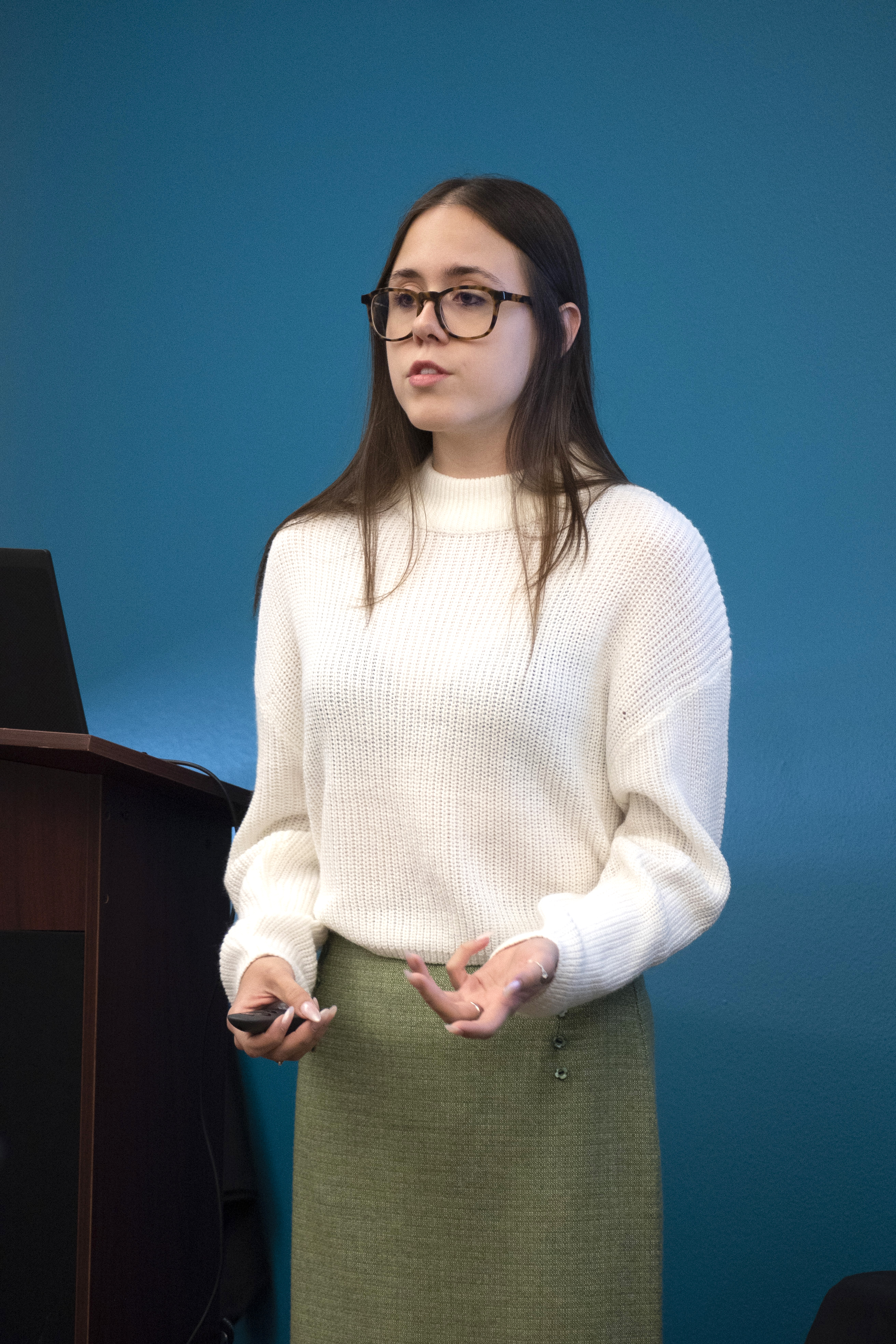 Photo of Natalie wearing a white sweater and a green skirt speaking.